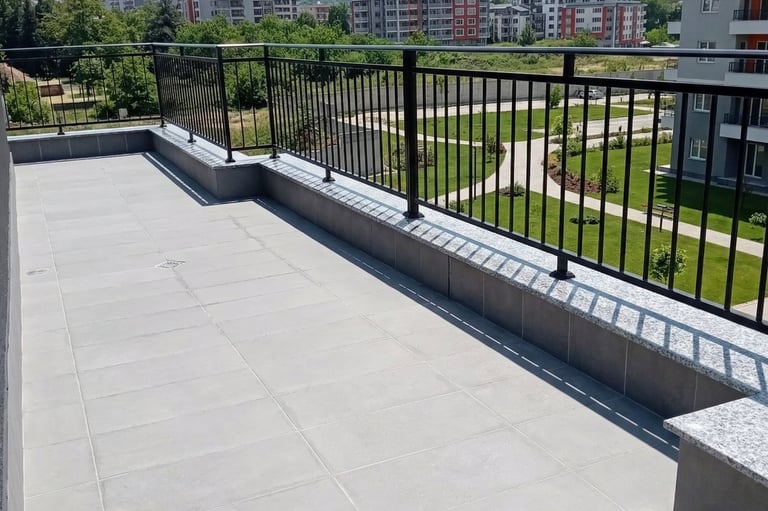Modern apartment balcony with grey tile flooring and black metal railing overlooking a landscaped park.