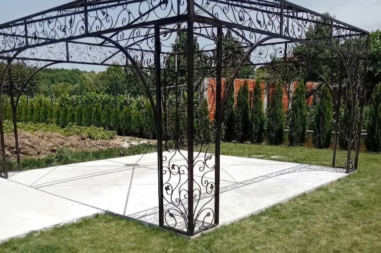 Ornate black wrought iron gazebo frame on a white concrete patio in a green garden landscape.