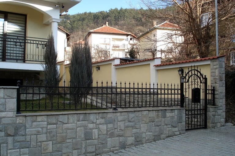 Luxury home with black wrought iron fence and stone wall masonry along a paved sidewalk.