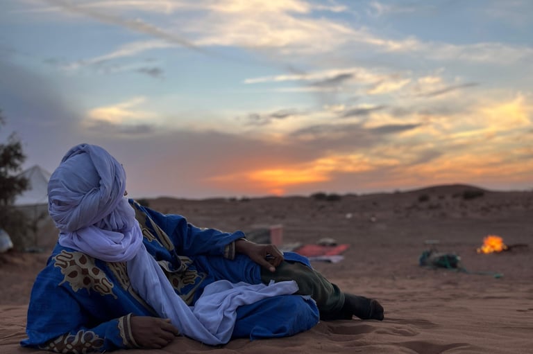 A man in traditional dress lies in the desert watching the sunset