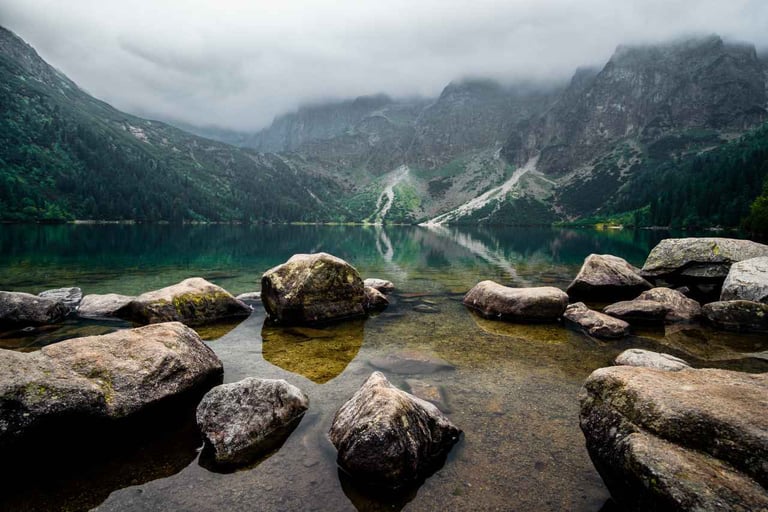 A calm, reflective mountain lake surrounded by rocks and partially shrouded in mist, with trees.