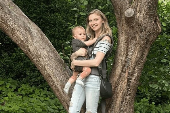Mother holding baby in a carrier by a tree, bonding in nature.