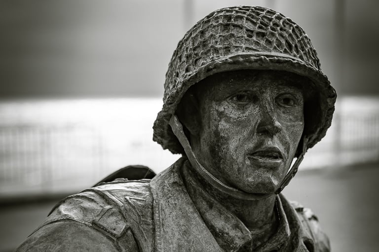 Gros plan sur le visage d'une statue de soldat en bronze, casque sur la tête, regard intense. Mer et ciel couvert en fond.
