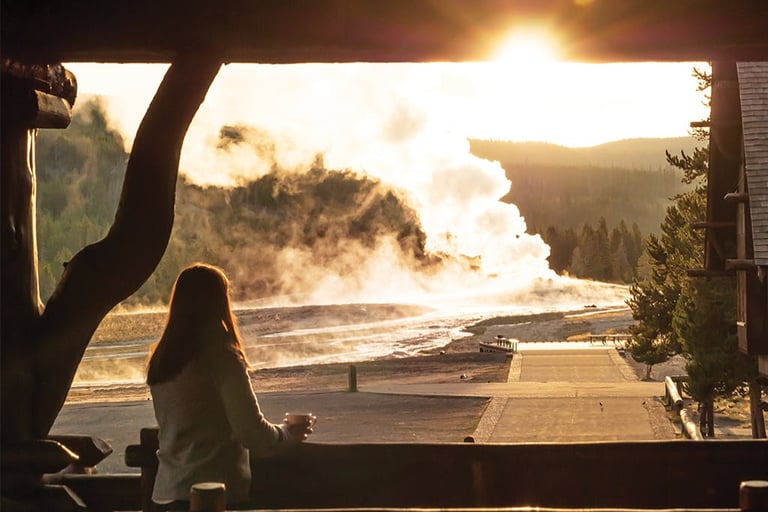 A woman watches the Old Faithful geyser erupt at sunset from a Yellowstone lodge porch.