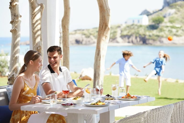 A couple enjoys a luxury outdoor lunch at a beach resort while children play on the grass.