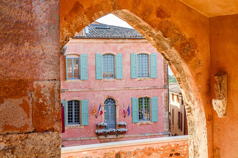 View of a pink Provencal building with teal shutters through a stone arch in Roussillon, France.