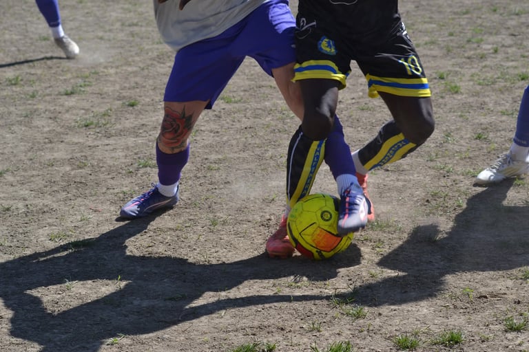Due calciatori lottano per il possesso di un pallone giallo-arancione su un campo in terra, con le g