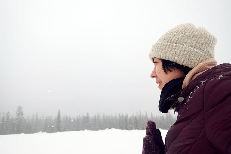 Person in winter jacket and knit hat standing in falling snow, looking across a snowy landscape with