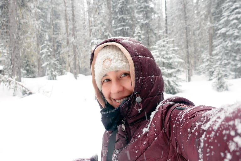 Person smiling during a snowy forest walk, wearing a winter jacket and hat with snow falling around.