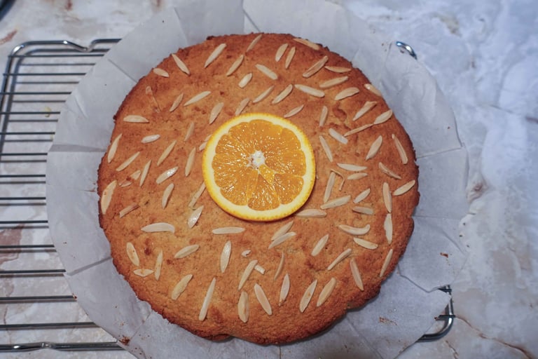 An almond orange cake topped with sliced almonds and an orange slice, cooling on a rack.