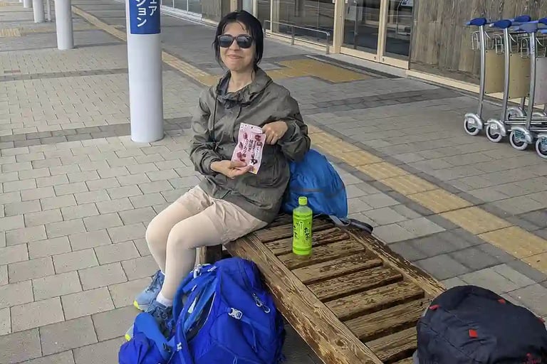 Woman sitting on a wooden bench outside an airport terminal with hiking backpacks and a bottle of green tea