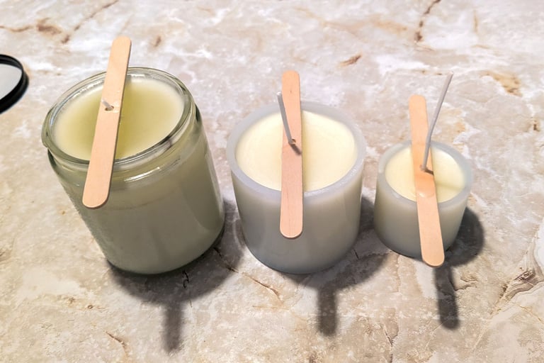 Three homemade candles cooling in glass containers on a marble surface with wooden sticks holding th