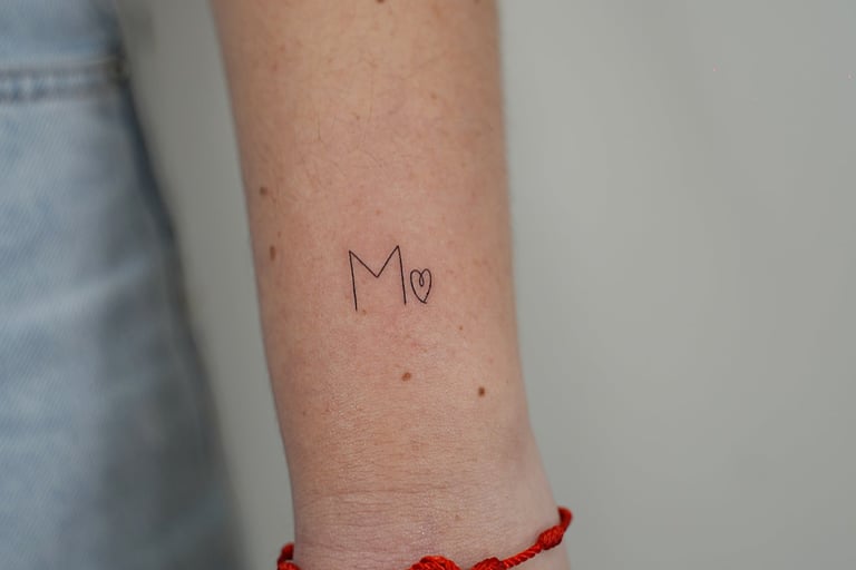 Tatuaje fineline en Madrid de inicial M con corazón pequeño en trazo fino en la muñeca