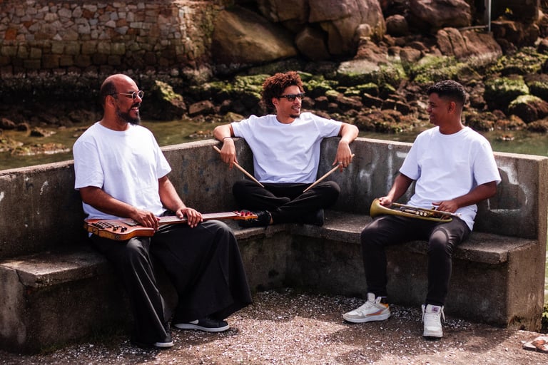 Three musicians holding instruments outdoors sitting on a stone bench by the water.