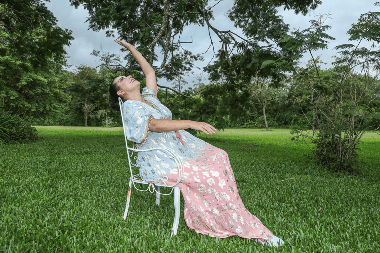 A woman in a floral dress leans back in a white garden chair in a lush green park.