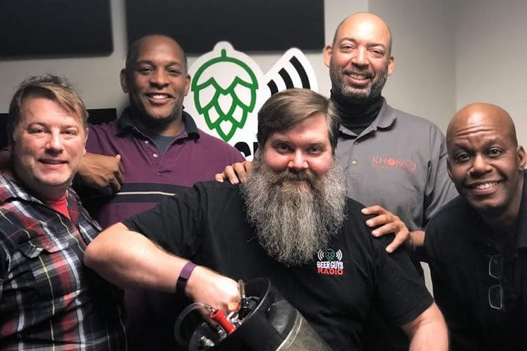 Five men posing for a photo at Beer Guys Radio with a beer keg and hop logo.