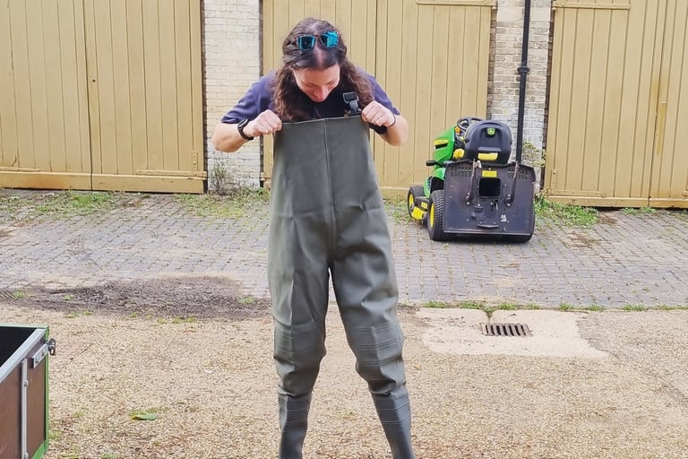 A woman putting on waterproof olive green fishing chest waders in a gravel courtyard.