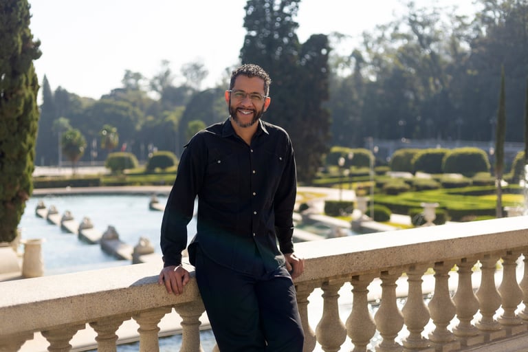 Foto do Mestre Mauricio apoiado em uma mureta, com um lago e um jardim ao fundo.