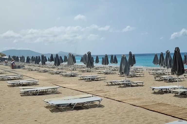 Empty sunbeds and closed grey umbrellas on a sandy tropical beach with blue ocean water.