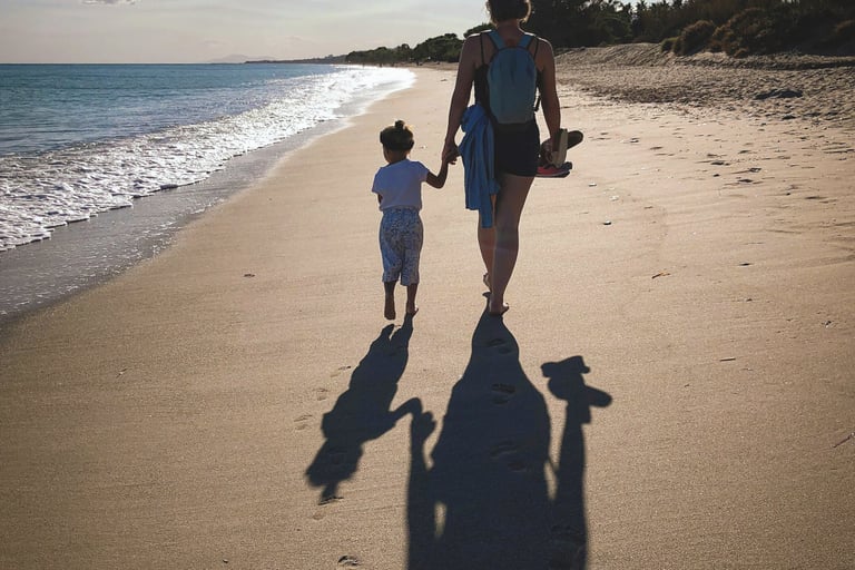 mother holds toddler's hand as they walk along a beach