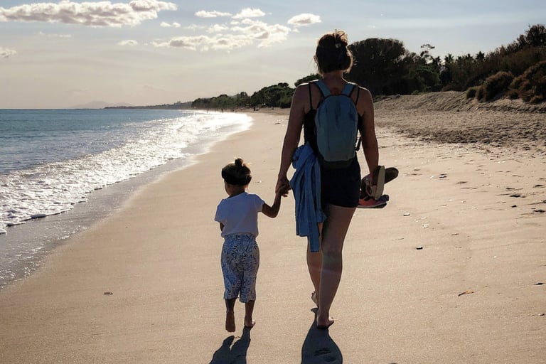 mother holds toddler's hand as they walk along a beach