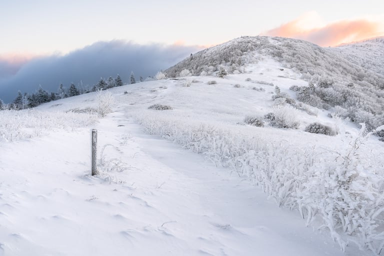 Snow-covered hiking trail leading up a frozen mountain ridge at sunrise with a cloudy sky.