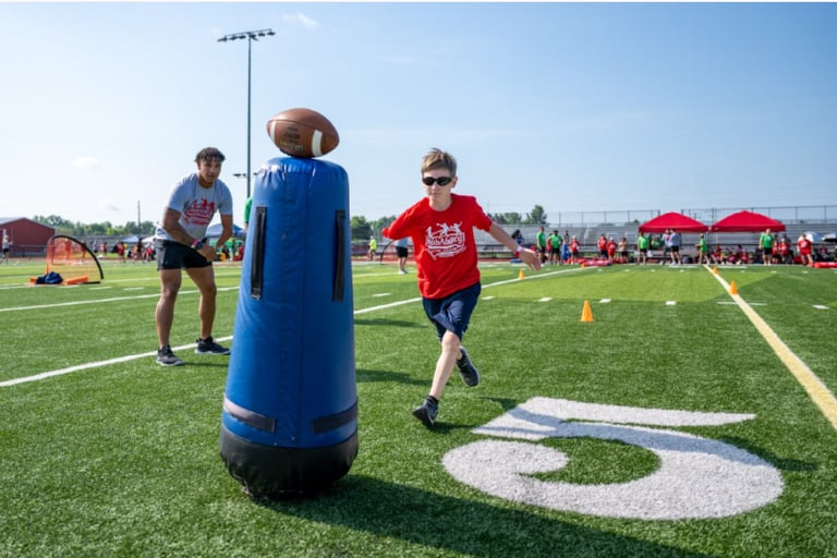 A young boy in a red t-shirt running through football drills with a coach on a turf field.