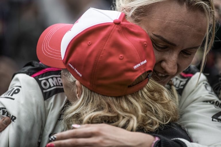 Two female race car drivers in OMP suits embrace to celebrate a victory on the track.
