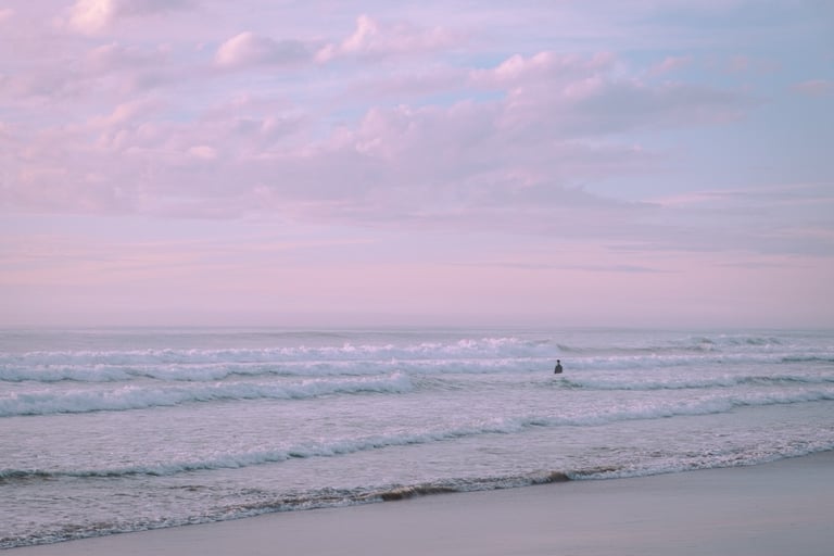 Serene shot of man in the distance standing in the ocean's waves. Pastel sunset sky.