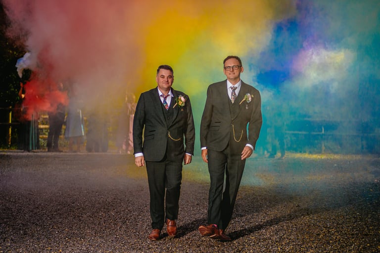 two grooms walking towards the camera with a rainbow of different coloured smoke bombs behind them