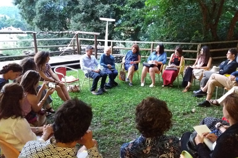 Cerchio di persone sedute all’aperto tra alberi e staccionata, in contesto informale