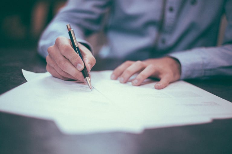 A person in a blue, button-up shirt sits at a desk writing on loose leaf paper