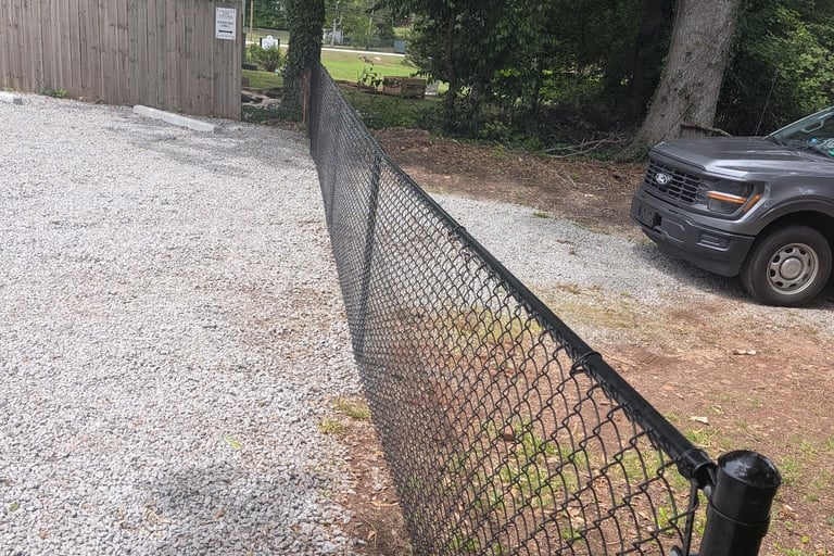 Black chain link fence separating a gravel driveway from a dirt path with trees and a grey pickup truck, enhancing security