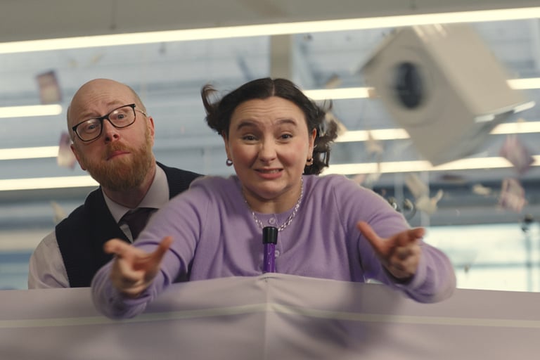 Woman in a lilac cardigan reaches forward as papers fly; man in glasses stands behind her.
