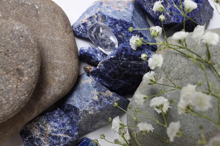 Cristales naturales de sodalita azul y piedras lisas de río acomodados junto a flores blancas. 