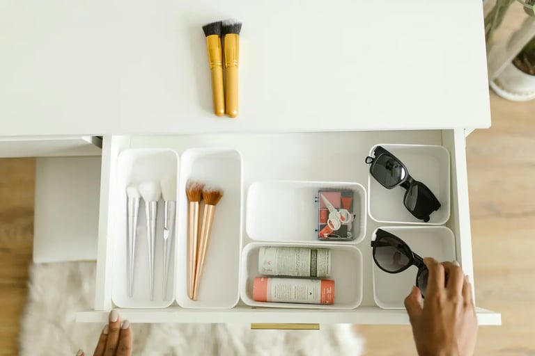 A person organizes makeup brushes and sunglasses in white plastic drawer dividers on a vanity.