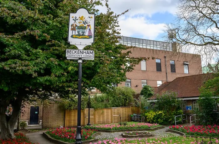 Beckenham Thornton's Corner sign standing in a public garden with flower beds and brick buildings.