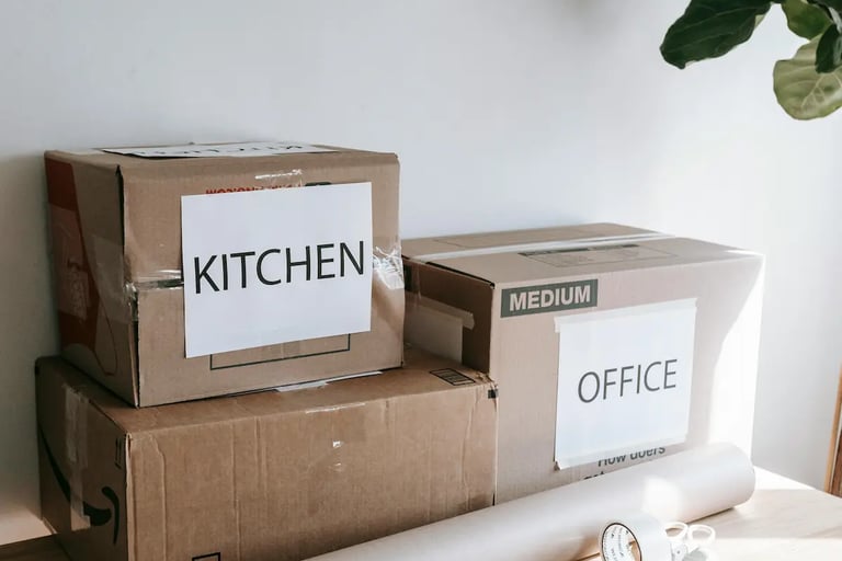 Cardboard moving boxes labeled Kitchen and Office on a table with packing paper and tape.