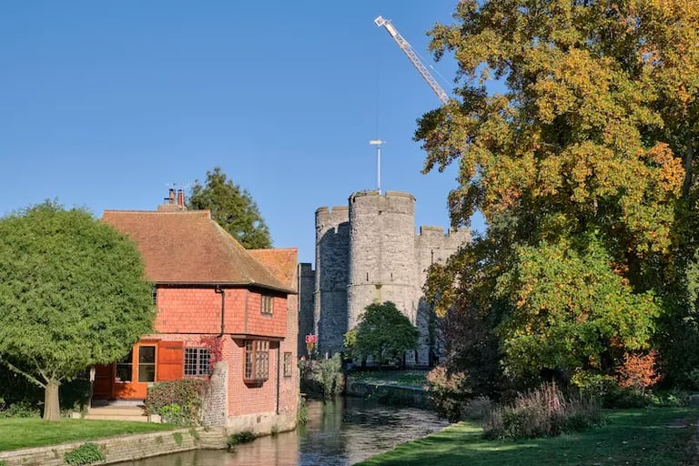 Historic Westgate Towers in Canterbury overlooking a river with autumn trees and a traditional brick house.