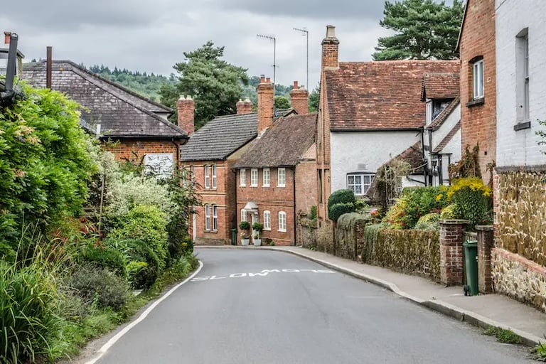 A narrow road winding through a charming English village with historic brick cottages and lush greenery.