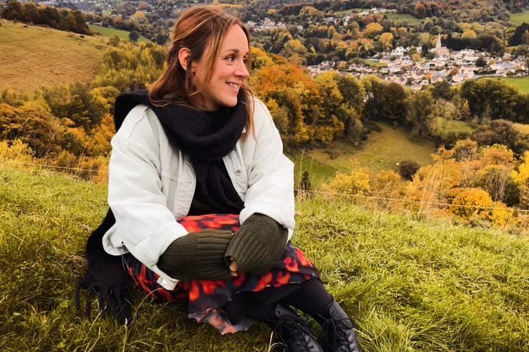 Smiling woman sitting on a grassy hill overlooking a scenic autumn valley with fall foliage and a small village.