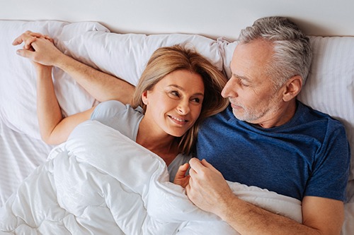 A smiling mature woman and man lying in bed holding hands and looking at each other affectionately.
