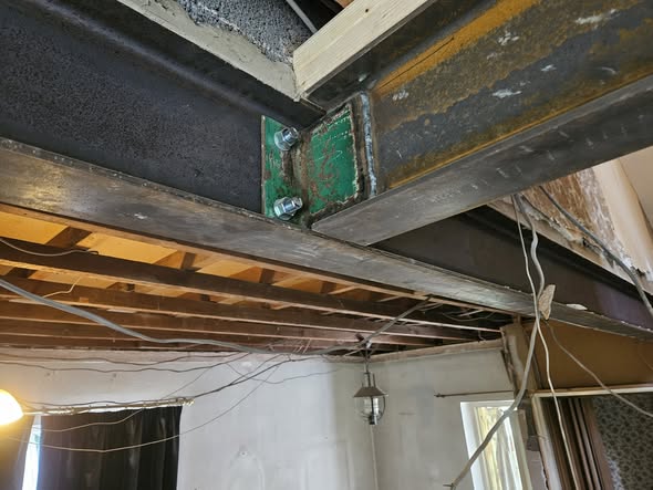 Structural steel I-beams joined by a green bolted splice plate during a home renovation.