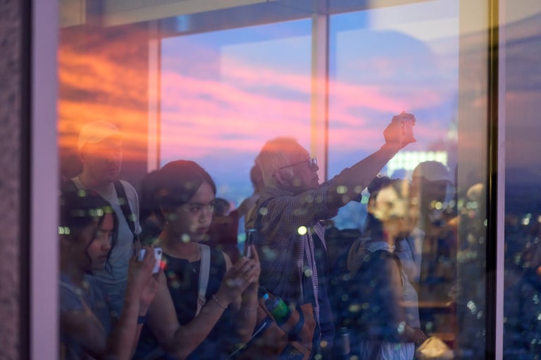 People taking photos of a colorful sunset from a glass observation deck overlooking a city skyline.