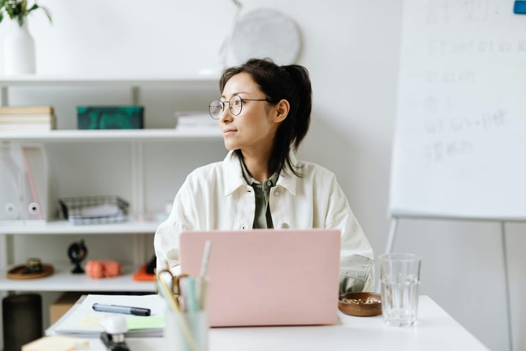 Professional woman with glasses working on a pink laptop in a modern, bright office setting.