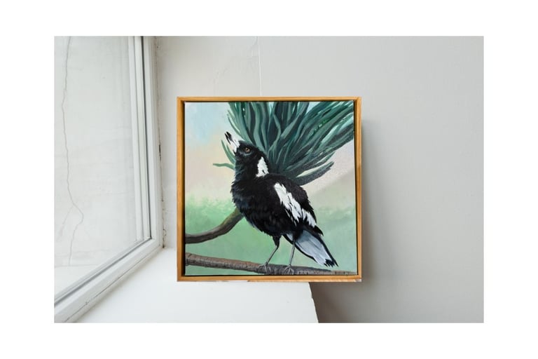 painting of an Australian magpie perched on a branch, framed in wood on a white windowsill.