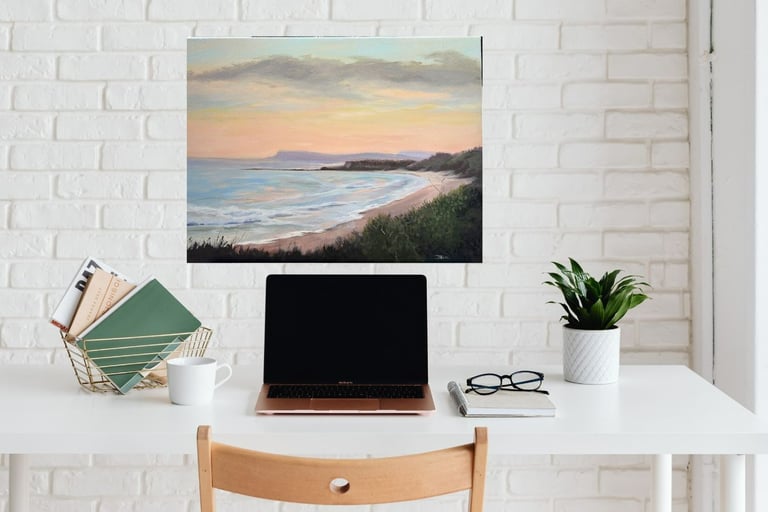  painting of a sunset beach hanging above a white desk with a laptop and a potted plant.