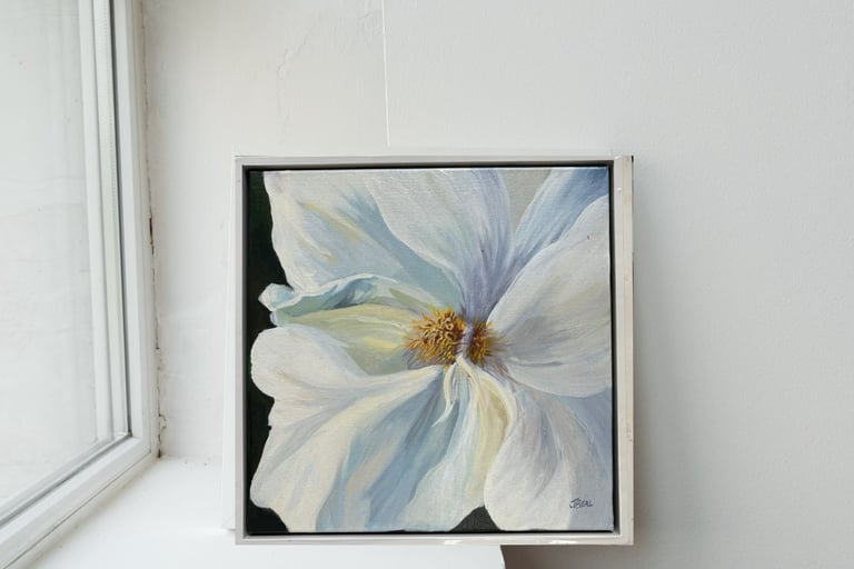 A square  painting of a white flower in a white frame on a windowsill.