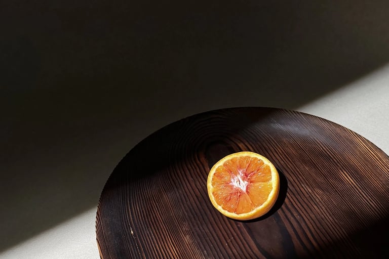 A single blood orange slice centered on a dark wooden platter under dramatic natural sunlight.