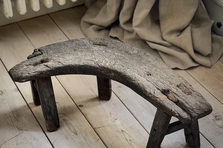 Rustic vintage curved wooden footstool on a light timber floor with linen curtains.